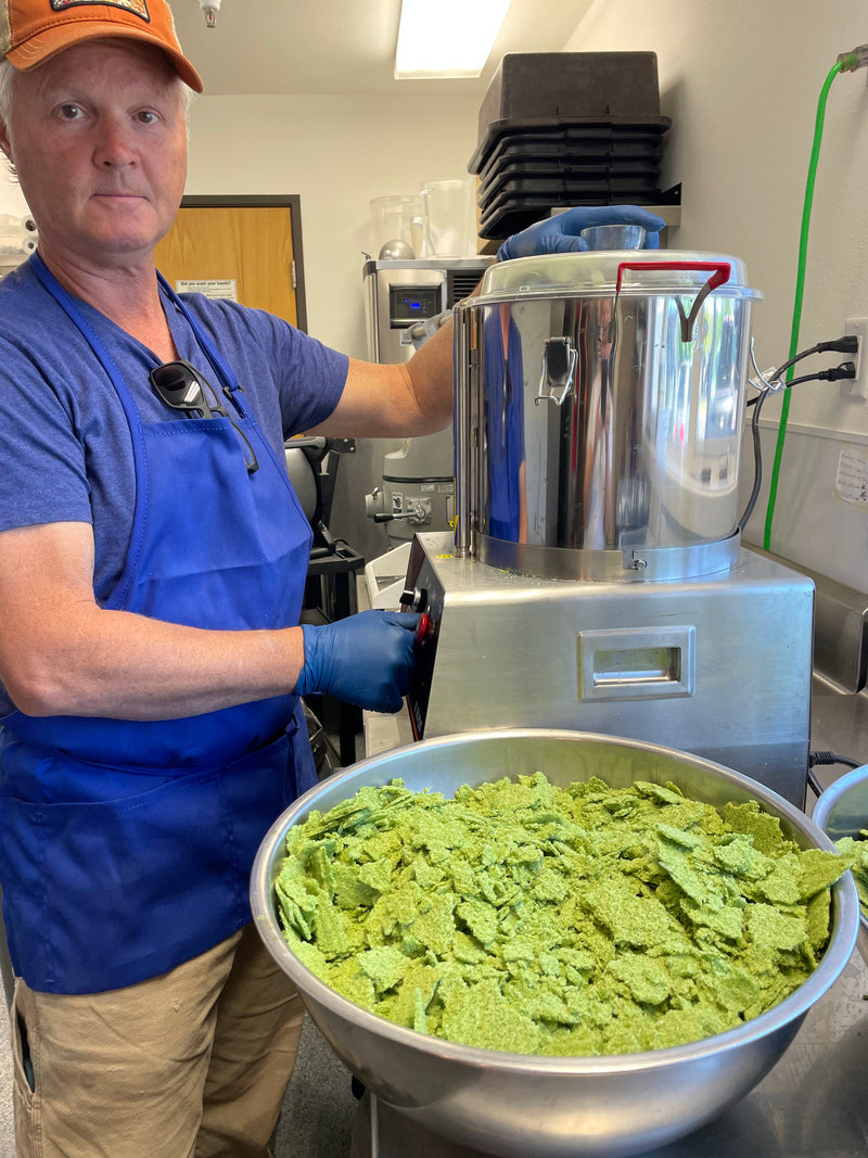 A man in a blue apron and orange cap operates a large stainless steel chip extruder, with a large bowl of green potato chips in the foreground.